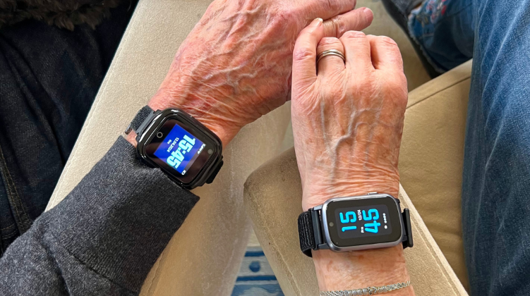 Två seniorer håller hand och båda bär Sensorems trygghetslarm. En av smartklockorna är av smal modell.